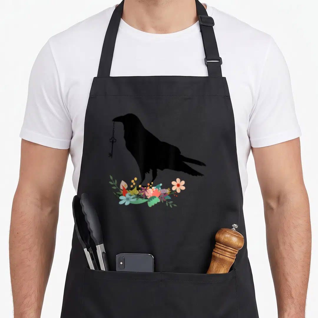 Blackbird crow bird apron - Black crow silhouette holding a key above green pink blue and orange flowers — shown on a black cotton canvas apron against a white background