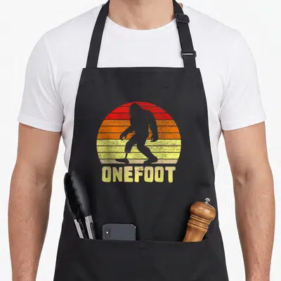 Onefoot bigfoot apron - Retro bigfoot silhouette with prosthetic leg against striped red orange yellow sun with ONEFOOT — shown on a black cotton canvas apron against a white background