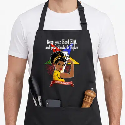 Star oes apron - Cartoon woman flexing arm in front of yellow red blue green star with Sisterhood banner — shown on a black cotton canvas apron against a white background