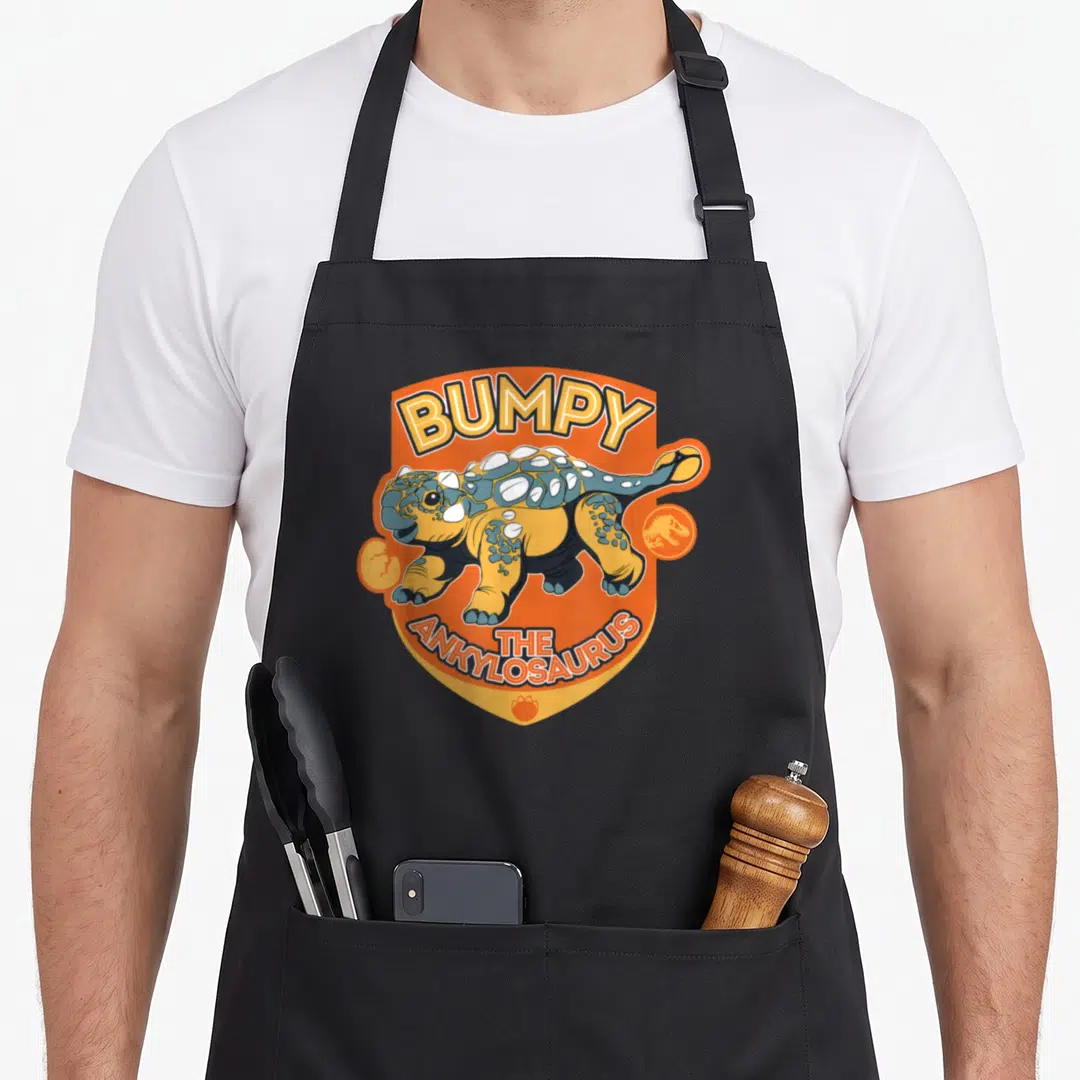 Jurassic ankylosaurus apron - Bumpy the Ankylosaurus cartoon on orange shield with footprint icons — shown on a black cotton canvas apron against a white background