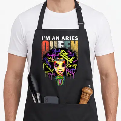 Ary queen birthday apron - I'M AN ARIES QUEEN text above cartoon woman with afro and dripping words Black Strong Proud — shown on a black cotton canvas apron against a white background