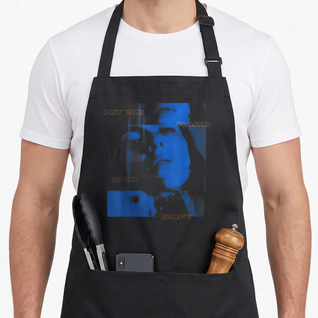 Billie hard apron - Blue woman's face with orange text HIT ME HARD AND SOFT on grey background — shown on a black cotton canvas apron against a white background