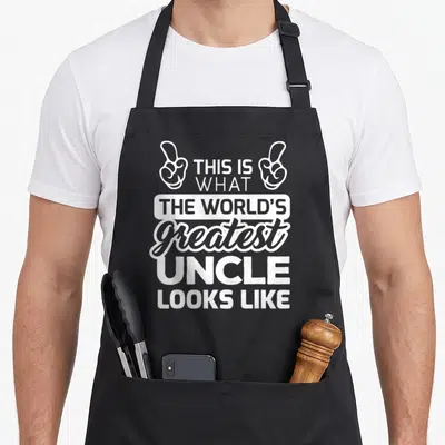 Uncle worlds greatest dad apron - White stacked text THIS IS WHAT THE WORLD'S greatest UNCLE LOOKS LIKE flanked by two — shown on a black cotton canvas apron against a white background