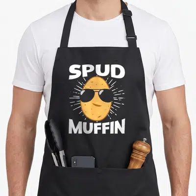 Spud plant based apron - Cartoon potato wearing sunglasses with SPUD MUFFIN text and white radiating lines — shown on a black cotton canvas apron against a white background