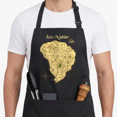 Isla worlds greatest apron - Retro tan map of Isla Nublar with labeled dinosaur zones and compass rose — shown on a black cotton canvas apron against a white background