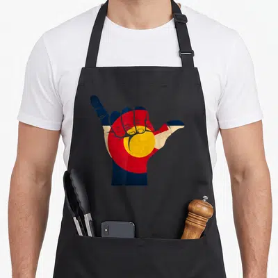 Shaka colorado apron - Hand making shaka gesture filled with blue red yellow and white flag pattern — shown on a black cotton canvas apron against a white background