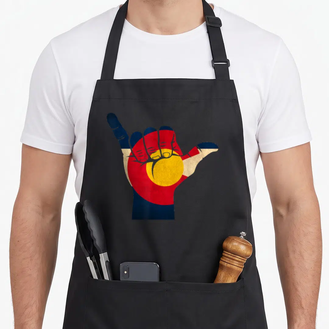 Shaka colorado apron - Hand making shaka gesture filled with blue red yellow and white flag pattern — shown on a black cotton canvas apron against a white background