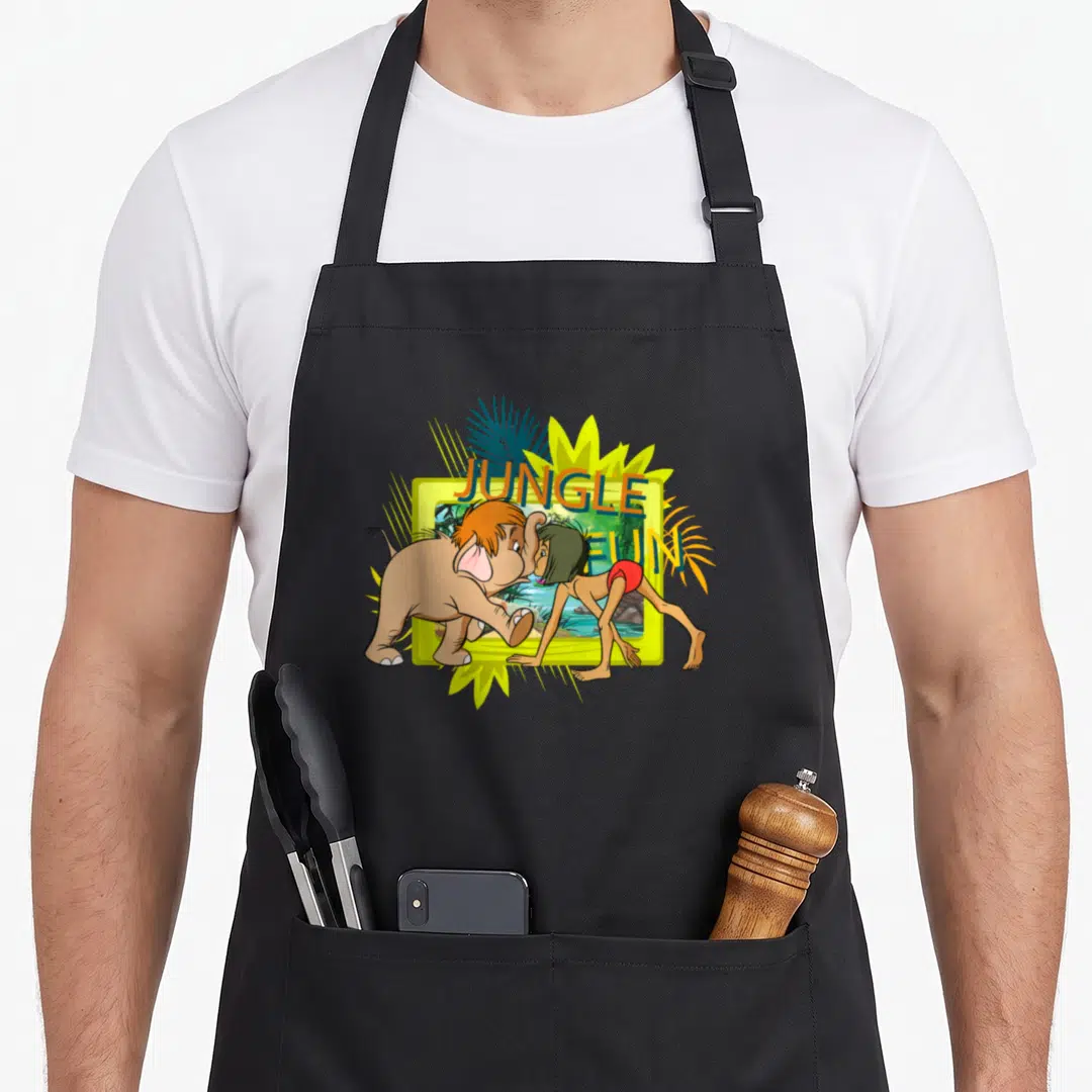 Book jungle juice apron - Cartoon elephant and boy facing each other beside yellow frame with JUNGLE FUN text in green — shown on a black cotton canvas apron against a white background