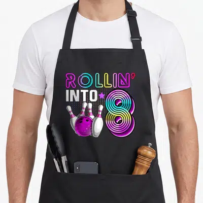 Rollin bowling birthday apron - Neon ROLLIN INTO 8 graphic with bowling ball and four pins in purple blue yellow — shown on a black cotton canvas apron against a white background