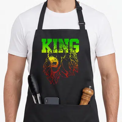 King lions apron - Cartoon lion head in green yellow and red with KING text above — shown on a black cotton canvas apron against a white background