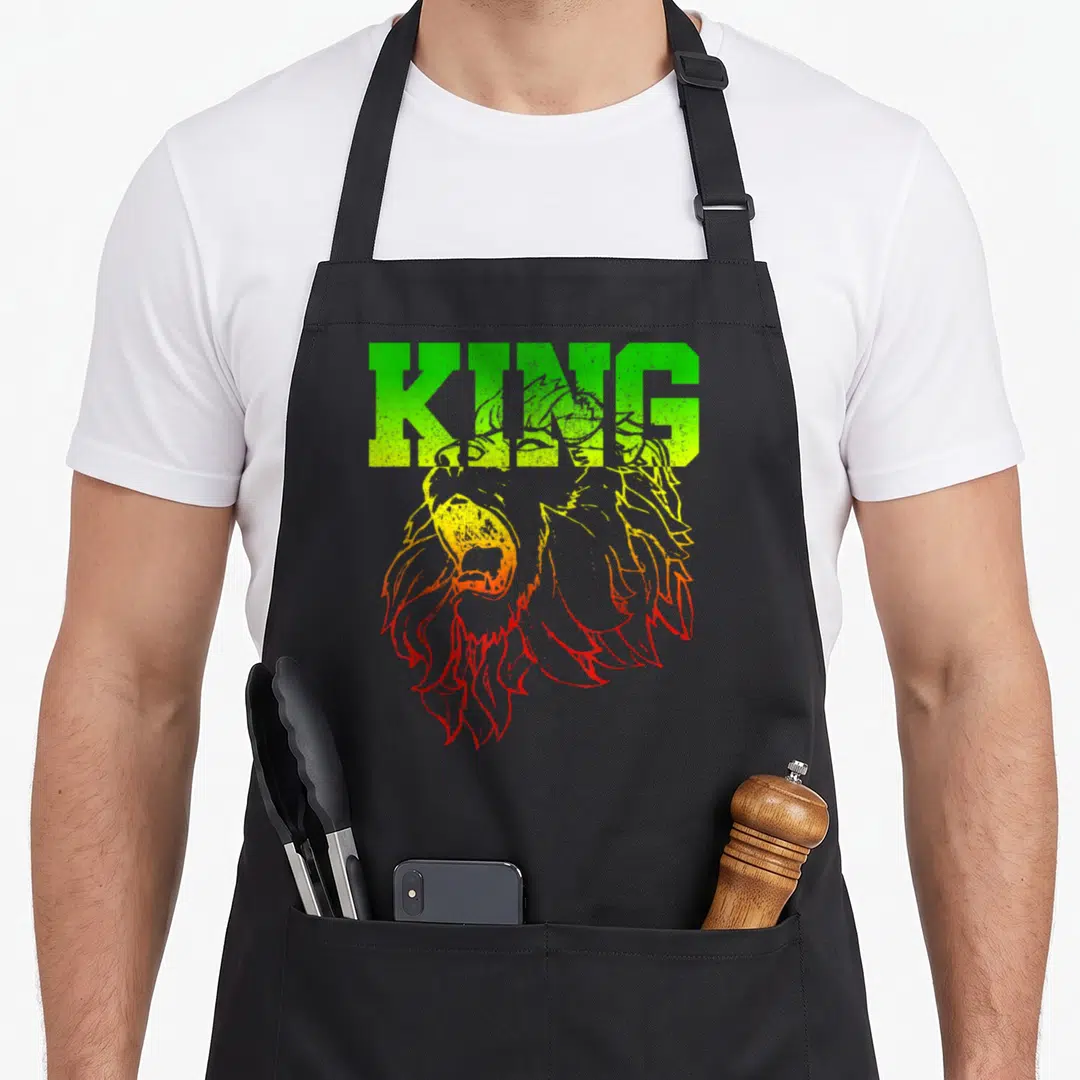 King lions apron - Cartoon lion head in green yellow and red with KING text above — shown on a black cotton canvas apron against a white background
