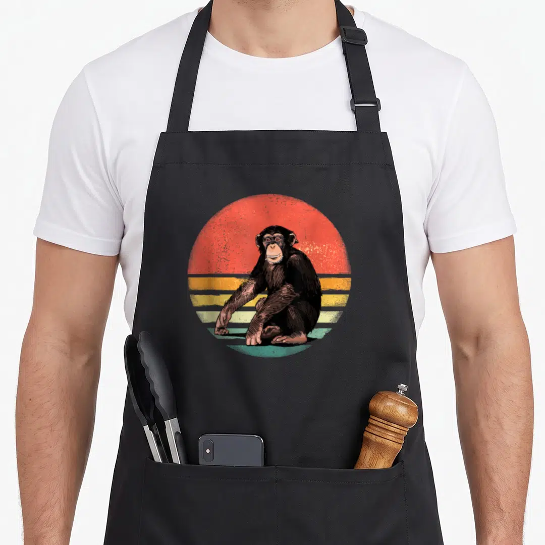 Chimpanzee animal apron - Seated chimpanzee in front of a red orange yellow and teal retro sunset circle — shown on a black cotton canvas apron against a white background
