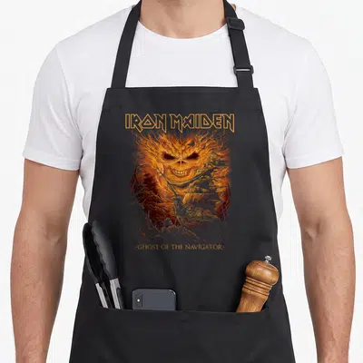 Ghost camping apron - Flaming skull and ship illustration with Iron Maiden text in orange and grey — shown on a black cotton canvas apron against a white background