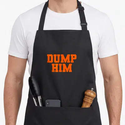 Dump crew apron - Orange block text reading DUMP HIM stacked in two lines — shown on a black cotton canvas apron against a white background