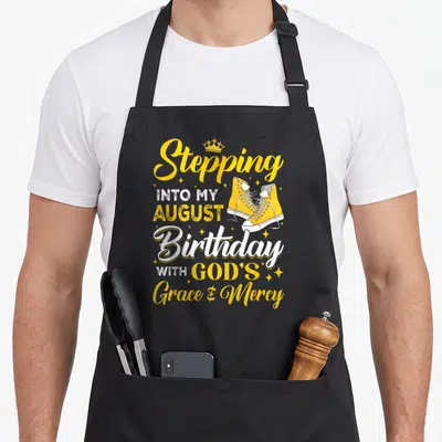 Stepping august birthday apron - Yellow and white text Stepping INTO MY AUGUST Birthday with sneakers and crown graphic — shown on a black cotton canvas apron against a white background