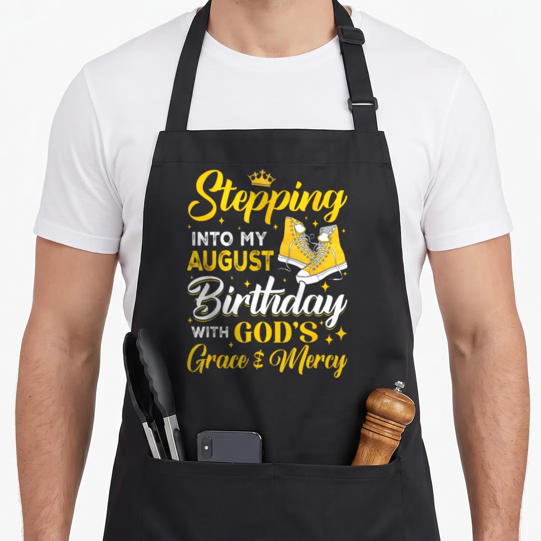 Stepping august birthday apron - Yellow and white text Stepping INTO MY AUGUST Birthday with sneakers and crown graphic — shown on a black cotton canvas apron against a white background