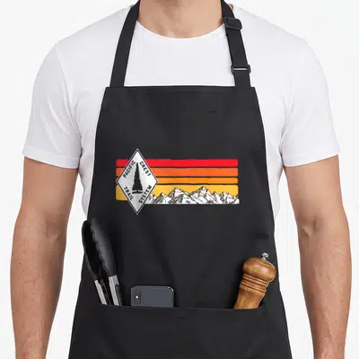 Marker pacific crest trail apron - Retro diamond logo with tree and mountain range over red orange yellow stripes — shown on a black cotton canvas apron against a white background