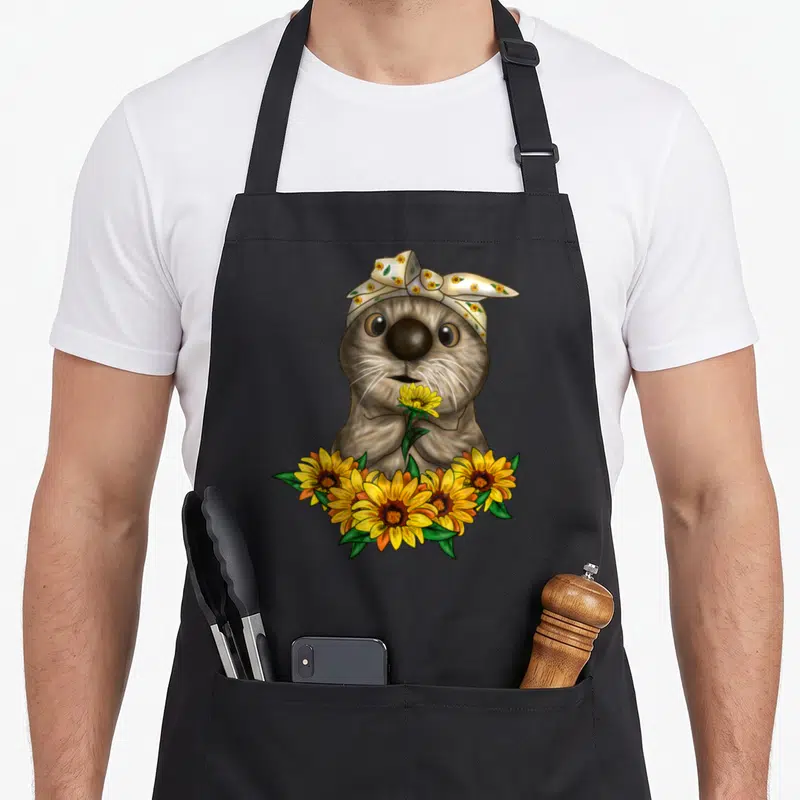 Otter sunflower apron - Cartoon brown otter with sunflower headscarf holding one yellow flower above a cluster of blooms — shown on a black cotton canvas apron against a white background