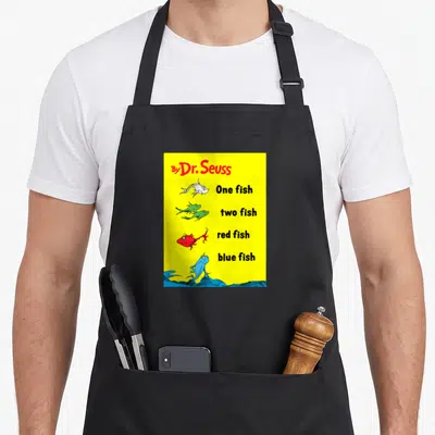 Book fish apron - Dr Seuss text and four colored fish on yellow background with blue waves — shown on a black cotton canvas apron against a white background