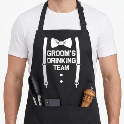 Drinking funny wedding aprons - White GROOM'S DRINKING TEAM text with bow tie and suspender straps ending in bottle — shown on a black cotton canvas apron against a white background