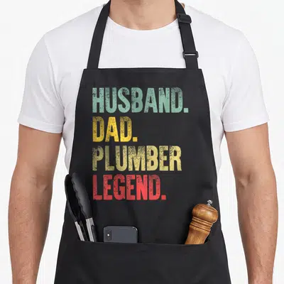 Plumber husband apron - Stacked HUSBAND. DAD. PLUMBER LEGEND. text in distressed green, yellow, and red block letters — shown on a black cotton canvas apron against a white background