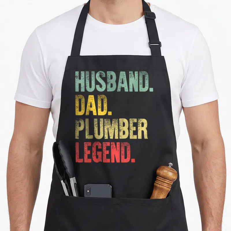 Plumber husband apron - Stacked HUSBAND. DAD. PLUMBER LEGEND. text in distressed green, yellow, and red block letters — shown on a black cotton canvas apron against a white background