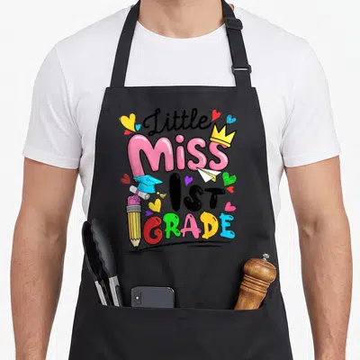 Cute 1st miss grade apron - Little Miss 1ST GRADE text with pencil, crown, and hearts in pink yellow blue green black — shown on a black cotton canvas apron against a white background