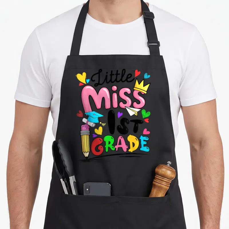 Cute 1st miss grade apron - Little Miss 1ST GRADE text with pencil, crown, and hearts in pink yellow blue green black — shown on a black cotton canvas apron against a white background