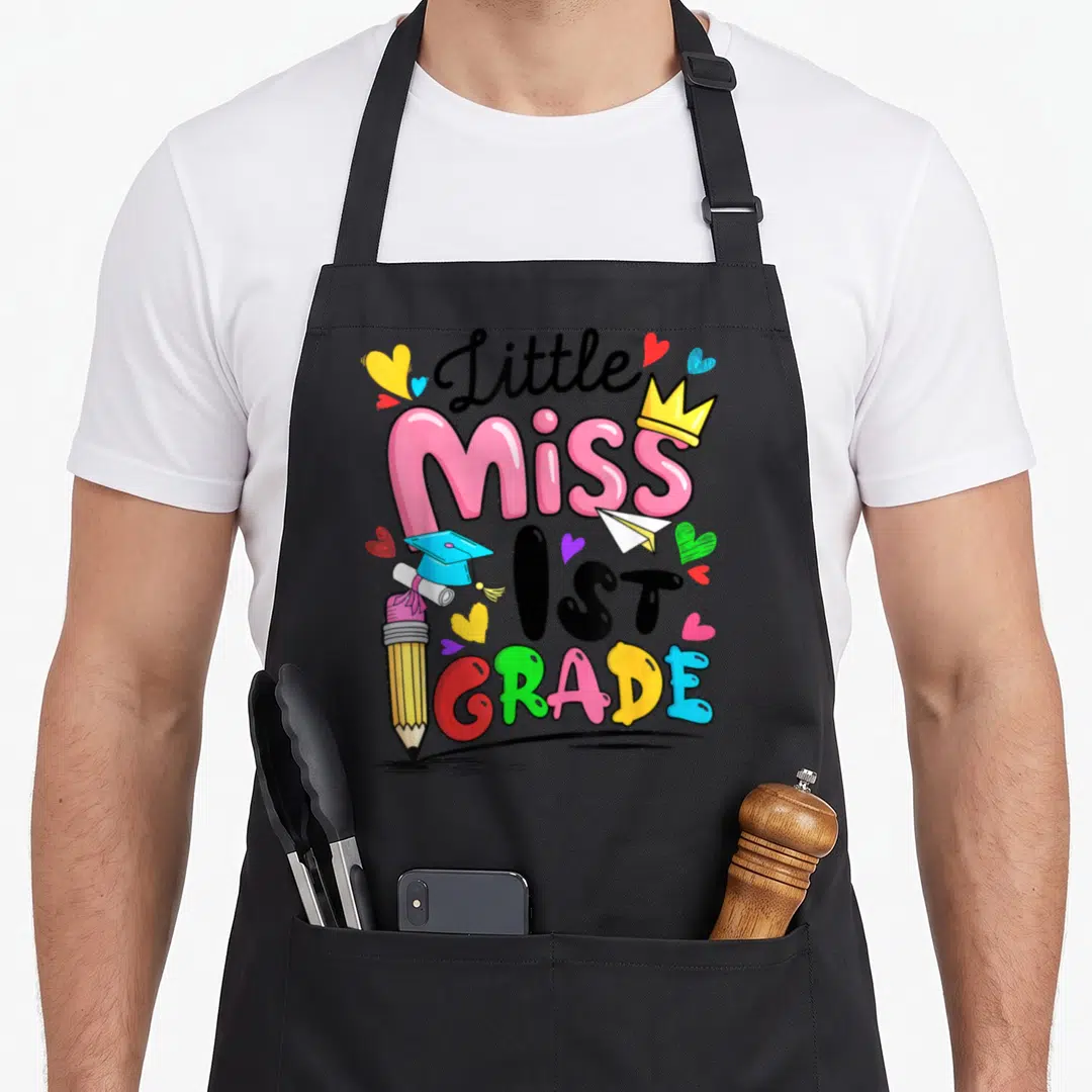 Cute 1st miss grade apron - Little Miss 1ST GRADE text with pencil, crown, and hearts in pink yellow blue green black — shown on a black cotton canvas apron against a white background