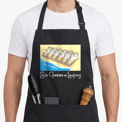 Geese christmas apron - Six Geese a Laying text below cartoon geese in sunglasses on beach chairs — shown on a black cotton canvas apron against a white background
