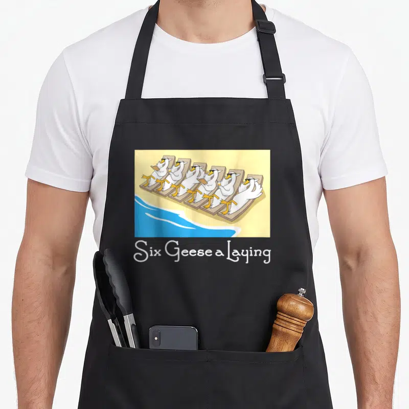 Geese christmas apron - Six Geese a Laying text below cartoon geese in sunglasses on beach chairs — shown on a black cotton canvas apron against a white background