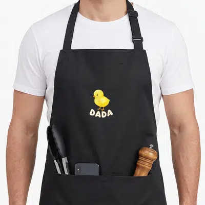 Dada chicken apron - Yellow watercolor chick illustration above off-white DADA text on black background — shown on a black cotton canvas apron against a white background
