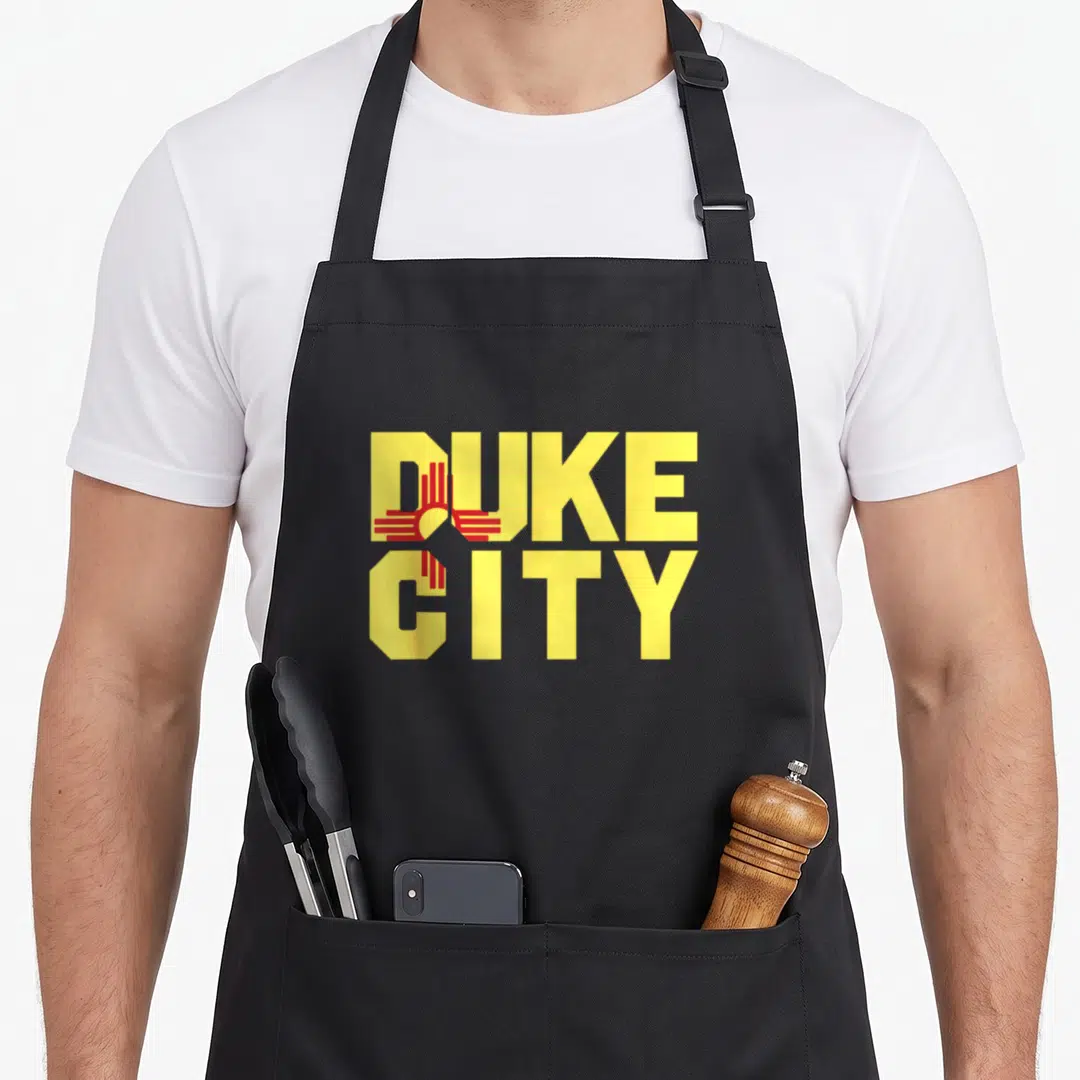 Funny duke mexico apron - Yellow DUKE CITY text with overlapping red Zia sun symbol representing New Mexico — shown on a black cotton canvas apron against a white background