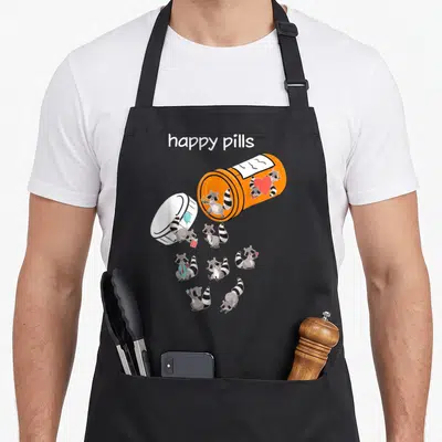 Pill funny raccoon lover apron - Orange pill bottle spilling cartoon raccoons holding balloons, gifts, and hearts — shown on a black cotton canvas apron against a white background