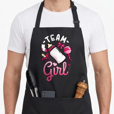 Retro gender reveal apron - Cartoon baby bottle surrounded by rattle, sock, pacifier, bow, and safety pin with TEAM — shown on a black cotton canvas apron against a white background