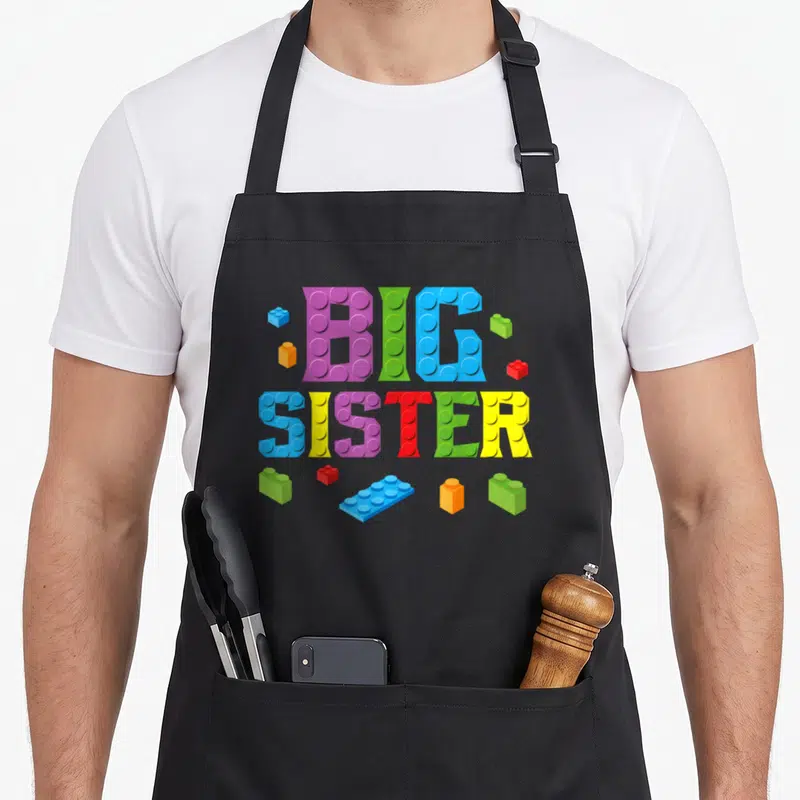 Sister grill master apron - BIG SISTER text formed by colorful toy bricks in purple green blue yellow red and orange — shown on a black cotton canvas apron against a white background