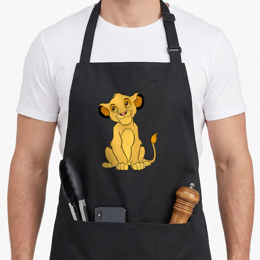 Cosplay lion apron - Seated cartoon lion cub in yellow and brown with pink nose centered on grey background — shown on a black cotton canvas apron against a white background
