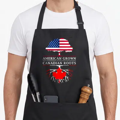 Grown canadian aprons - Tree split by text with US flag canopy and Canadian maple leaf roots in red white blue — shown on a black cotton canvas apron against a white background