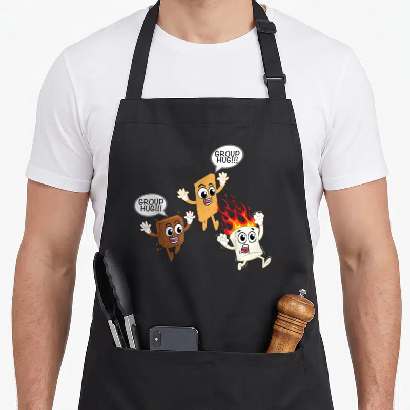 Roasted camping apron - Cartoon s'mores characters chasing a flaming marshmallow with group hug speech bubbles — shown on a black cotton canvas apron against a white background