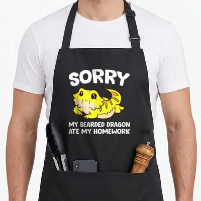 Bearded ate dragon apron - Yellow cartoon bearded dragon with white text SORRY MY BEARDED DRAGON ATE MY HOMEWORK — shown on a black cotton canvas apron against a white background