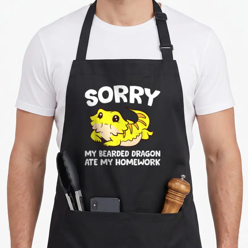 Bearded ate dragon apron - Yellow cartoon bearded dragon with white text SORRY MY BEARDED DRAGON ATE MY HOMEWORK — shown on a black cotton canvas apron against a white background