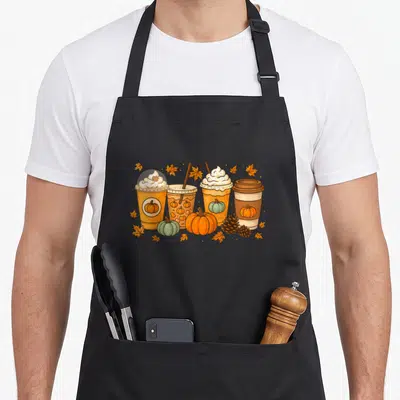 Coffee pumpkin halloween apron - Four cartoon pumpkin spice latte cups with pumpkins and autumn leaves in orange brown — shown on a black cotton canvas apron against a white background