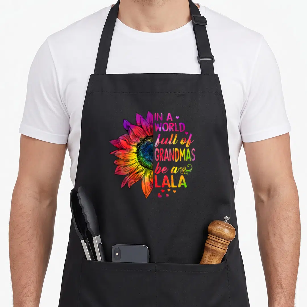 Lala grandma's aprons - Rainbow watercolor half sunflower next to stacked text IN A WORLD full of GRANDMAS be a LALA — shown on a black cotton canvas apron against a white background