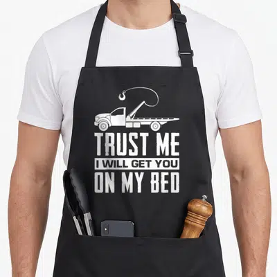 Trust truck apron - White tow truck with raised hook and distressed text TRUST ME I WILL GET YOU ON MY BED — shown on a black cotton canvas apron against a white background