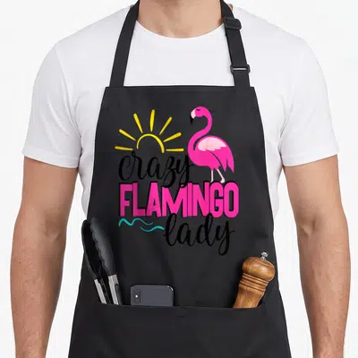 Cruise pink flamingo apron - Cartoon pink flamingo with yellow sun and stacked crazy FLAMINGO lady text — shown on a black cotton canvas apron against a white background