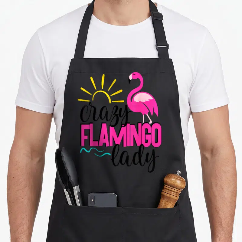 Cruise pink flamingo apron - Cartoon pink flamingo with yellow sun and stacked crazy FLAMINGO lady text — shown on a black cotton canvas apron against a white background