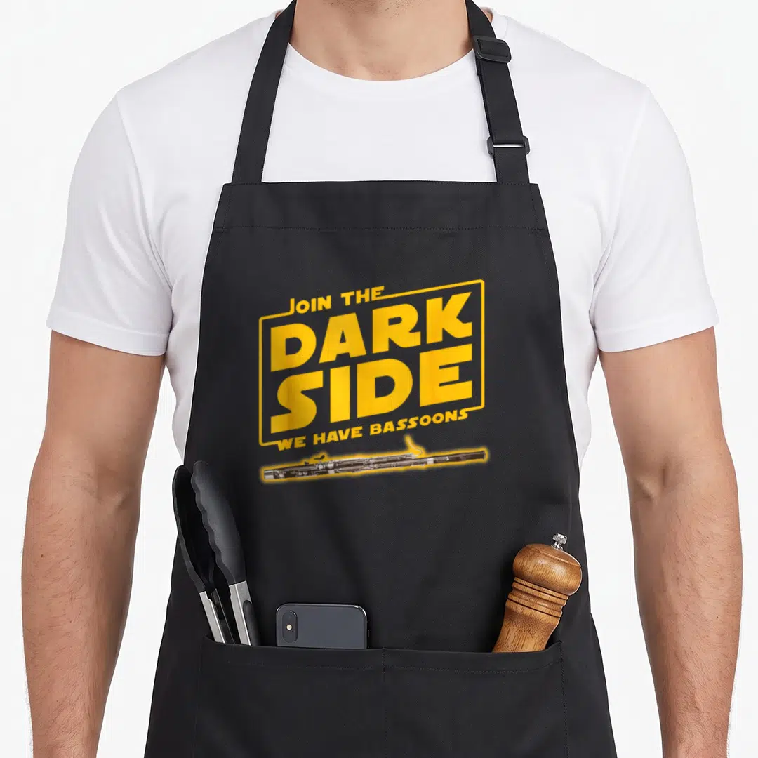 Join side addict apron - Yellow framed text Join The Dark Side We Have Bassoons above horizontal black bassoon — shown on a black cotton canvas apron against a white background