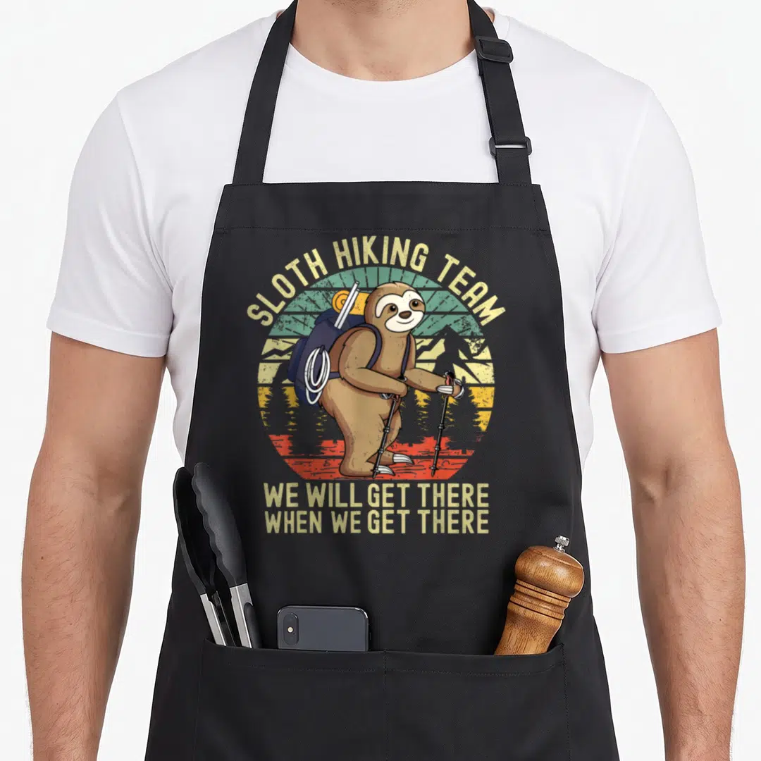 Well sloth apron - Retro brown sloth with blue backpack and beige text reading Sloth Hiking Team We Will Get There When — shown on a black cotton canvas apron against a white background