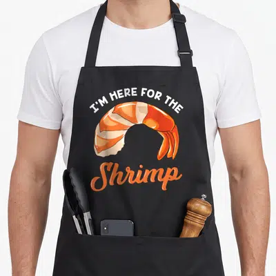 Seafood crazy shrimp man apron - Cartoon orange shrimp with I'M HERE FOR THE Shrimp text in white and orange script — shown on a black cotton canvas apron against a white background