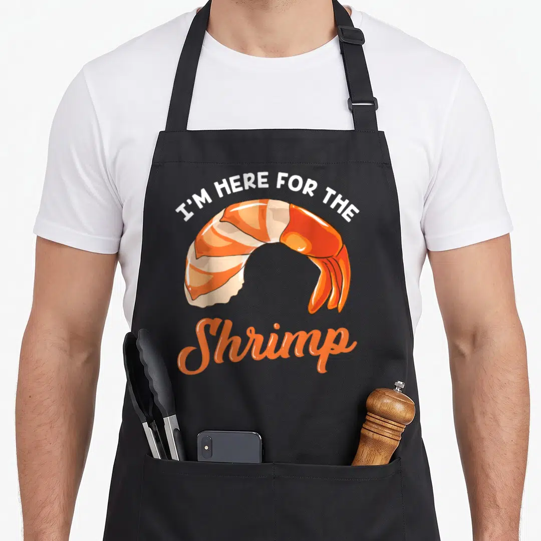 Seafood crazy shrimp man apron - Cartoon orange shrimp with I'M HERE FOR THE Shrimp text in white and orange script — shown on a black cotton canvas apron against a white background